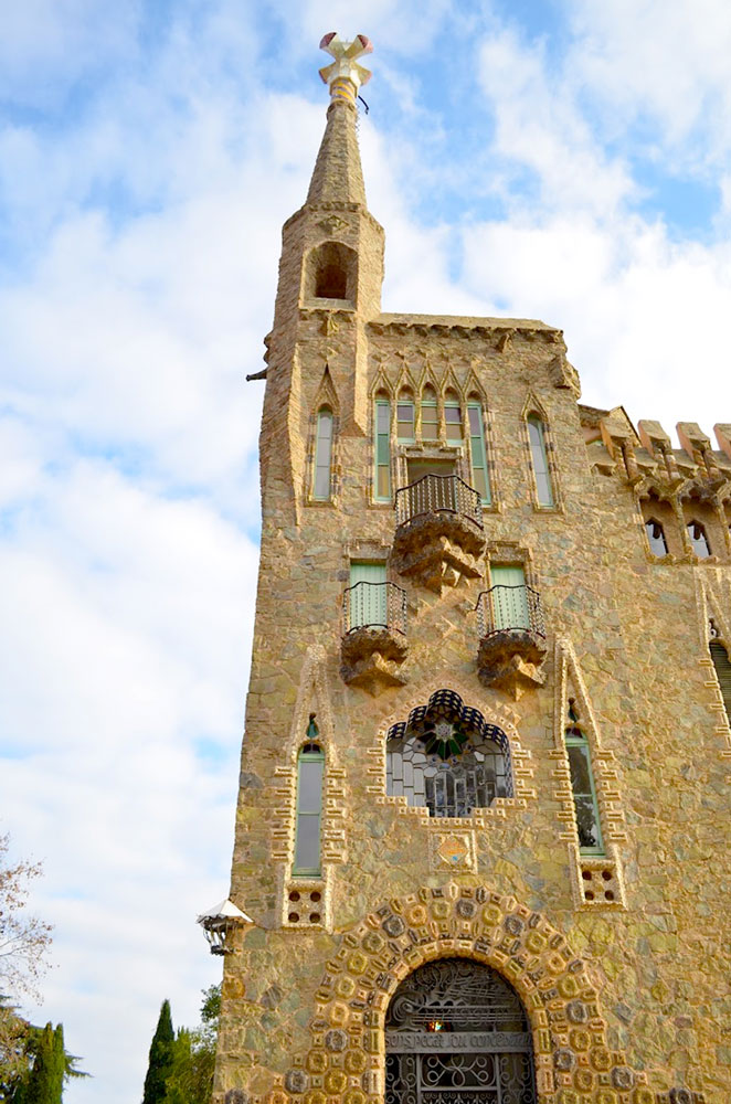 the tall stone tower of torres bellesguard designed by gaudi stands very tall. There's intricate mosaics throughout the entire face, with a cross at the top, and a few balconies on the side more layers to the facade, 