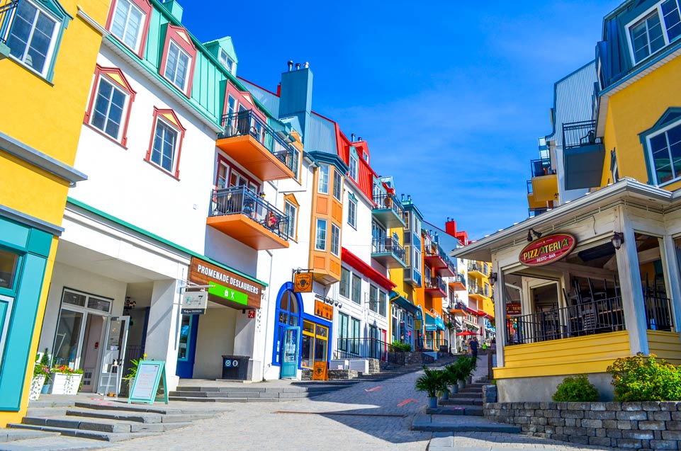 the view of the main pedestrian walking street in mont tremblant village. The image is bright and colourful because the sky is blue without a cloud in the sky. The buildings are each a unique fun painted coloured. On the corner is a large covered terrace with the sign for Pizzateria.