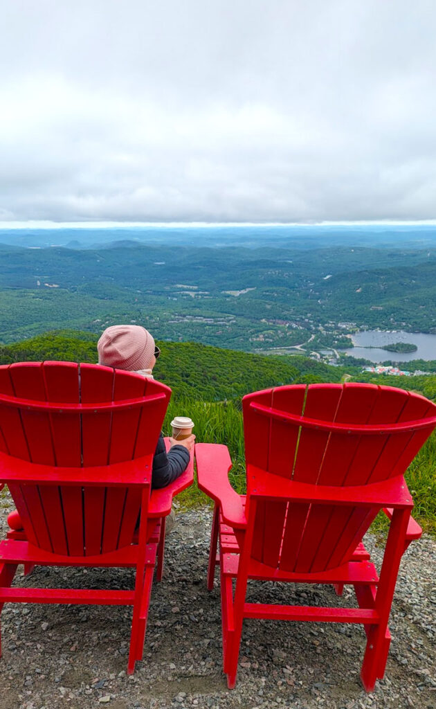 behind two large red chairs. A woman is sitting on the left one, with a cup of tea and shes wearing a pink winter hat. She's looking out onto the mountain and lakes that are way below. The sky is overcast, but you can see that it clears up in the distance.