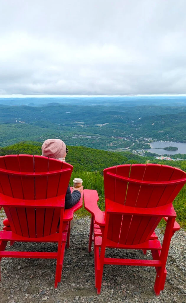 behind two large red chairs. A woman is sitting on the left one, with a cup of tea and shes wearing a pink winter hat. She's looking out onto the mountain and lakes that are way below. The sky is overcast, but you can see that it clears up in the distance.