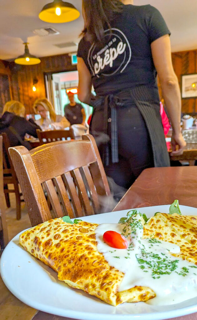 a delicious looking savoury crepe, that looks more like an omelette. There's a white sauce with a tomato on top. Its on a wooden table. Blurred in the background is a wait staff with her back to the camera, shes all in black but her t shirt clearly has the logo and it says Crepe