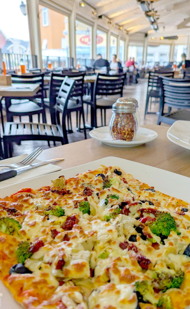 a large squared thin crust pizza that's loaded with vegetables and cheese on top taking up the bottom third of the image. In the background, blurred, is the restaurant with empty tables and chairs, and an awning shielding the terrace from the beating sun.