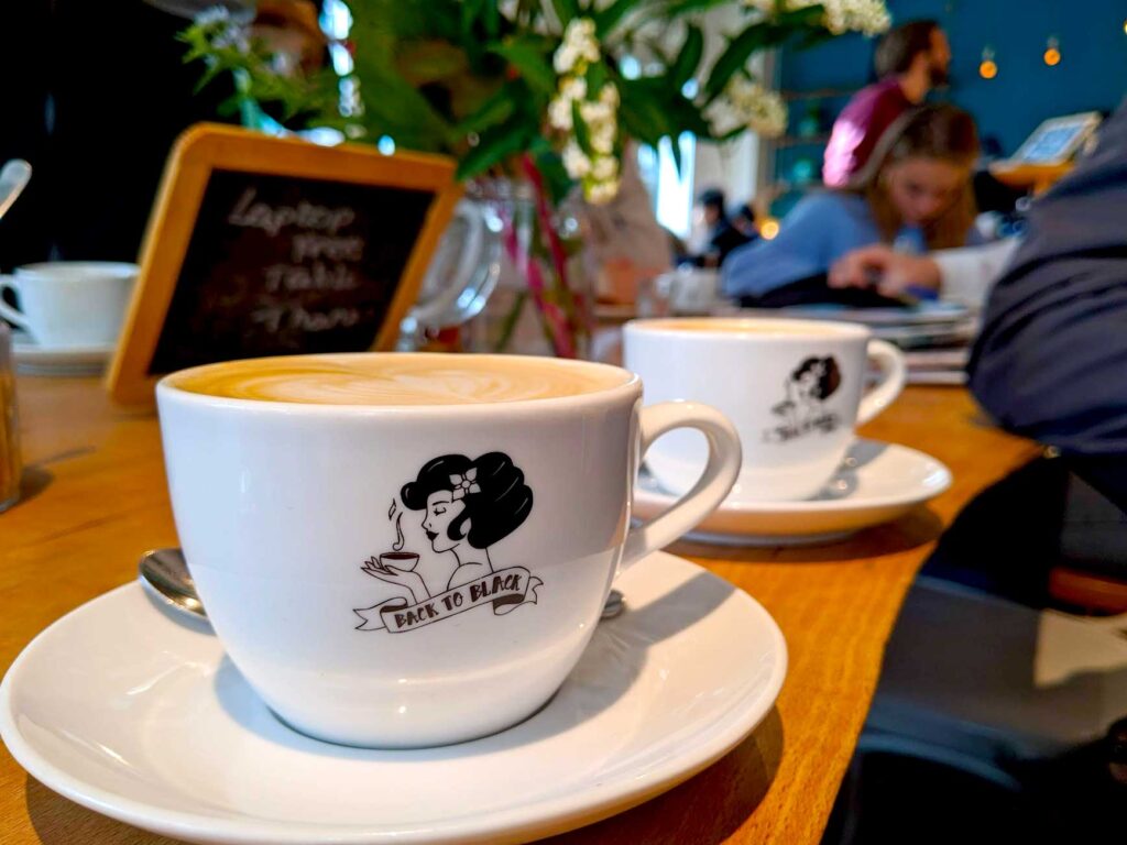 back to black coffee in amsterdam. Sitting at a long communal table, other people are blurred in the background, doing work. There are two cappucino mugs in focus at the front. The logo on the white mug is black and white. the profile of a woman holds a warm mug with steam coming out of it, she has a short black bob with a flower in her hair. There's a banner underneath wit Back to Black