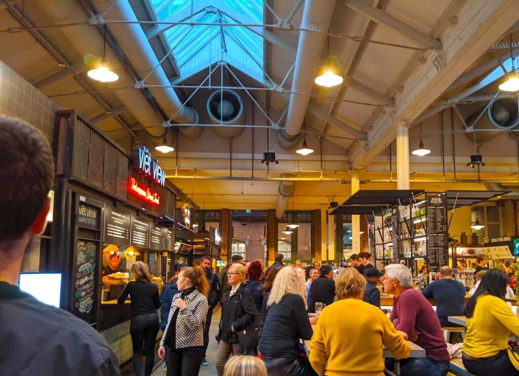 inside foodhallen with bustling crowds and food vendor stalls. There are three restaurants in a row on the left side, people are standing in line, walking towards it, or sitting at one of the many tables in the middle.