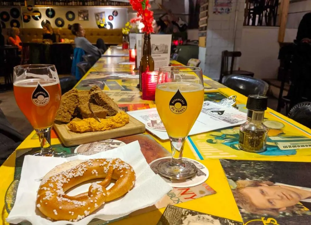 inside de prael brewery in amsterdam. A long table going all the way to the other side of the restaurant. There's only one other group sitting at the table, far on the opposite side.  We've ordered some bread with hummus, there are two beers, and a giant preztel. The lighting is dim in the brewery. 