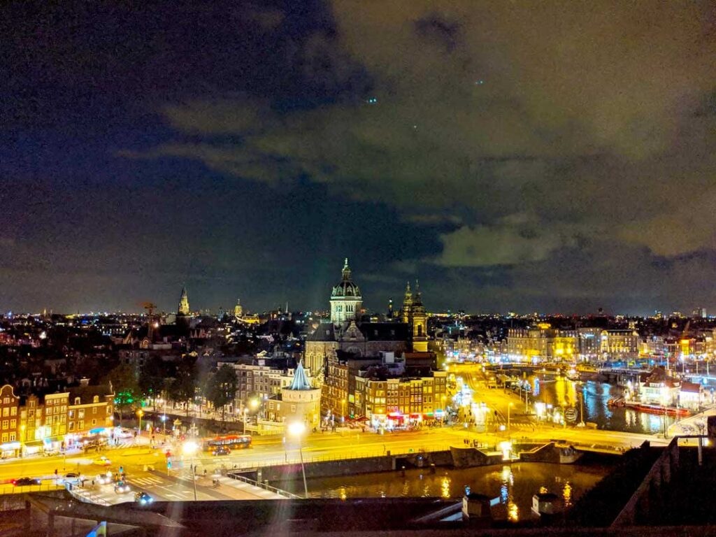 the view from the skylounge double tree amsterdam eats. The top half of the photo is all sky, you can see a dark grey cloud coming in on the right of the photo. Then the city is in night mide below, the lights give the city a vibrant yellow colour. You can see red kights from street signs, traffic lights, and cars and trams driving through the streets.