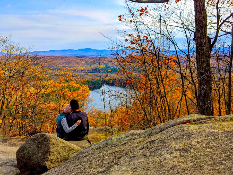 a couple sitting on a rocky mountain top. They are hugging each other wearing fall jackets. There are fall coloured trees all around them (orange, red, and yellow), and below there's a lake. Off into the distance you can see the continuation of the mountain range, but it's blending in to the blue sky