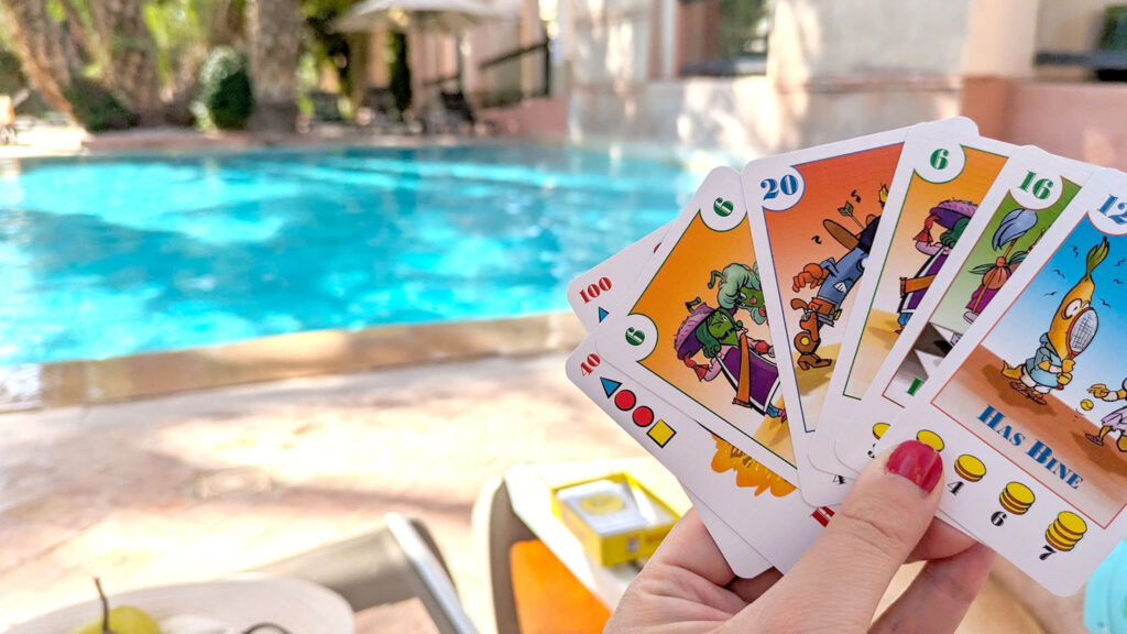 a woman holding five cards in her hands, displaying them so she can see what she's holding. They are fun comic cards that have cartoon beans on them, each one is a different number and there are coin amounts underneath the artwork. Behind the cards, blurred in the background is a bright blue pool. 