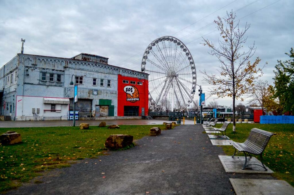 a walkway on a rainy gloomy day in the old port leads your way to a warehouse looking building, that doesn't look like it's in the best state. A quarter of the building is painted in bright red, with the logo for sos labrynthe on it. Behind the building is a large ferris wheel. 