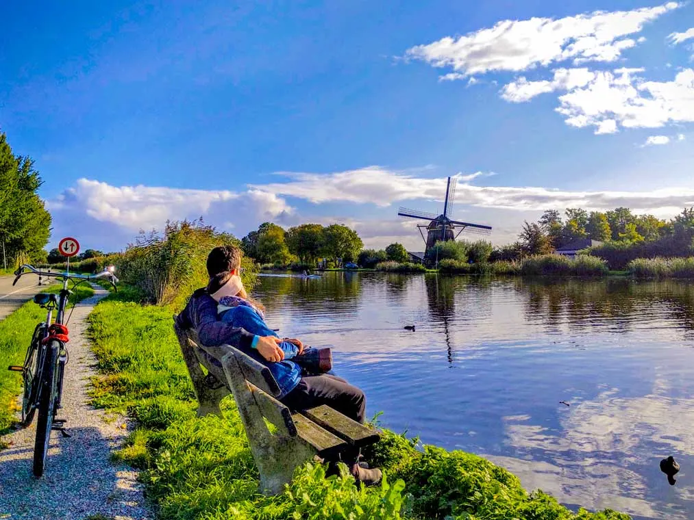 a couple sits on a bench, his arms wrapped around her shoulders. They overlook a calm canal with an old windmill across from it, it's reflection in the water. 
There's a bike leaning on it's stand behind the couple - their mode of transportation. Now they relax and enjoy the view. 