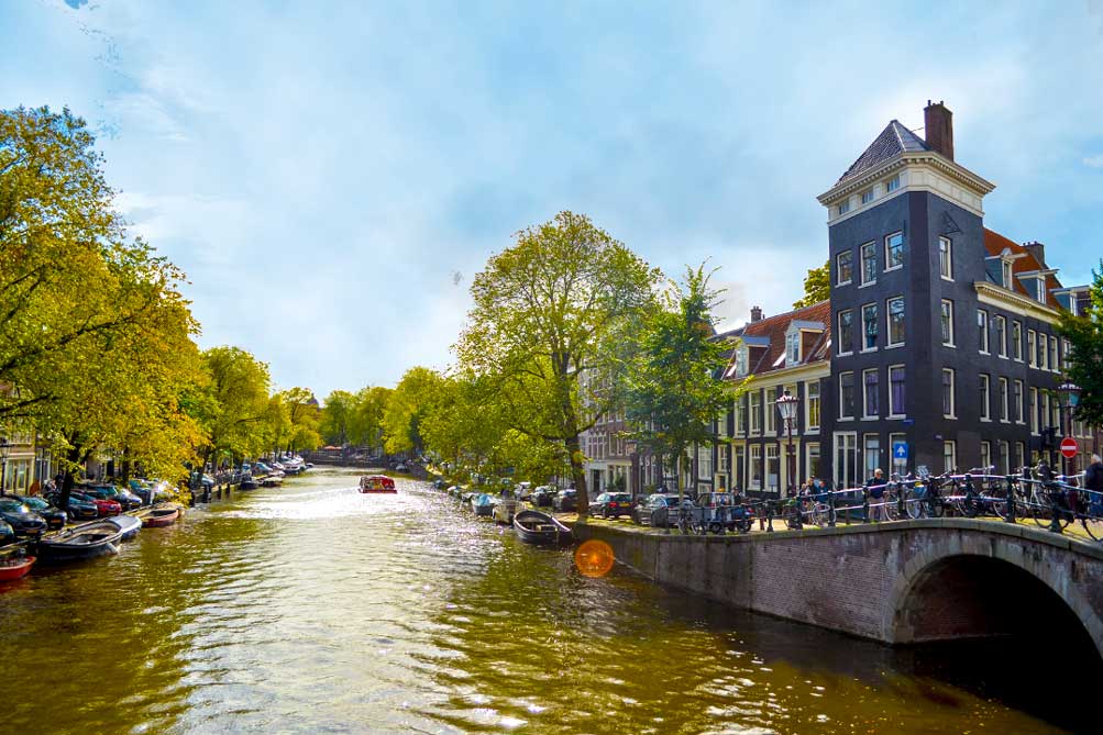 amsterdam canals on a sunny day. The lush green trees line the canal, wondering if the trees are rubbing off on the canal water... it's also looking quite green. 
Boats are docked in the water, cars are parked on the street, and bikes are locked up on the bridge railing on the right of the picture. You only get a peak of a amsterdam canal house as theres only the corner without any trees to block the view. 