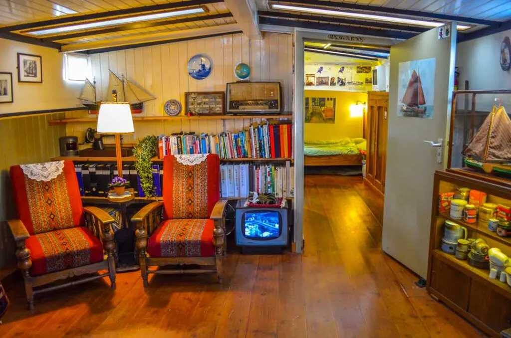 on board a house boat in amsterdam. It's narrow, wood floors and looks like the decor hasn't been updated in years. It is quite nautical feeling with white wood panneling and ship decor throughout. There are two old timey chairs in the corner, with a filled bookshelf behind them and it looks like a mini tv from the 50s on the floor.