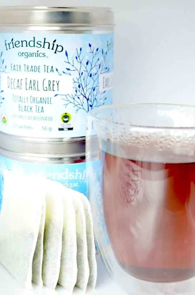 a double walled mug sits in the foreground of the image, with a dark rich reddish brew inside. It's steaming hot. There are standing unmarked blank tea bags all crunched together, but still standing upgrght next to the mug. they're probably also leaning on the stacked tins behind them. You can't see the bottom tin, but it matches the branding of the top one. Which has a bright blue paper wrapped around it. It says decaf earl grey tea 