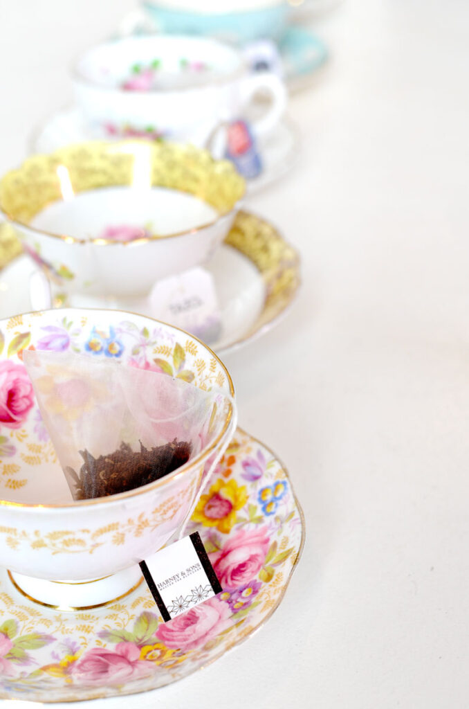 a lineaup of teacups go down in a vertical line across a white table. EAch one is different, and filled with a different tea bag. They are waiting to be filled with water and steeped for a tasting. The colours of the teacups all match with white, but their accent colours still compliment each other. From pink, to yellow, and a bright blue