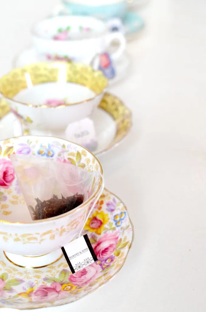 a lineaup of teacups go down in a vertical line across a white table. EAch one is different, and filled with a different tea bag. They are waiting to be filled with water and steeped for a tasting. The colours of the teacups all match with white, but their accent colours still compliment each other. From pink, to yellow, and a bright blue