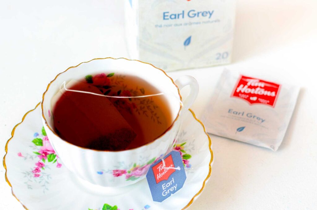 Tim hortons earl grey tea in a tea cup with the tea bag on the side. The tea cup is mostly white with some pink floral accents and gold rims. You can see the tea bag label that rests on the saucer, there's a red tim hortons logo, and below it says earl grey. There's the individual bag packaging laying on the table, and the full box behind it. 