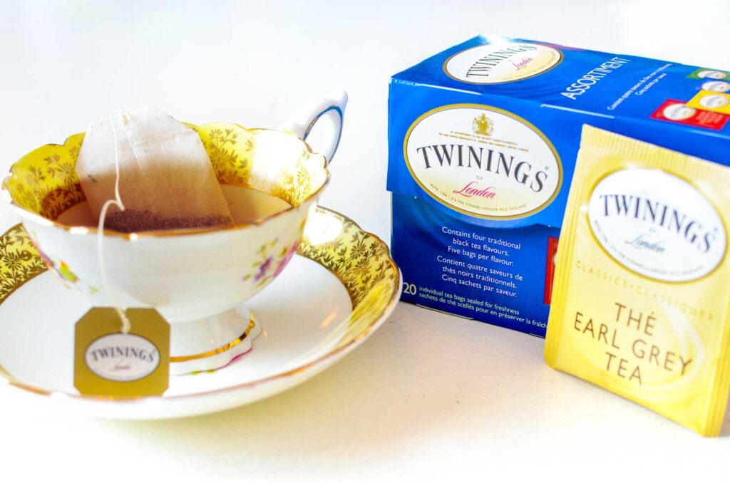 a delicate white teacup sits in a white saucer. The teacup and saucer match with yellow and gold patterned rims. The teacup seems like it was made to match the Twinings packaging. A tea bag sits, steeping in the cup. The string is attached to a gold label outside with the logo for twinings. Next to the teacup is a blue box for twinings tea bags, and outside, leaning against the box is the individual packaging for Twinings earl grey tea. It's a yellow package, with gold accents, and a white label for the logo. 