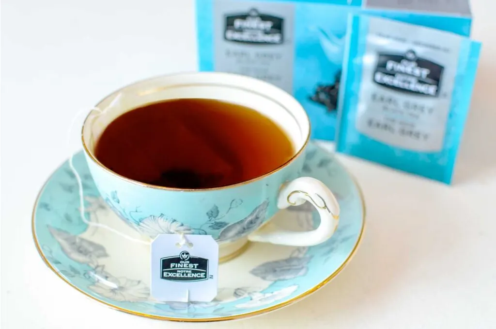 a light blue tea cup and saucer, filled with a dark brew. A label attached to the string shows that it's brewing our finest excellence tea. Slightly blurred in the background is the blue and silver box and individual packaging for the tea. 