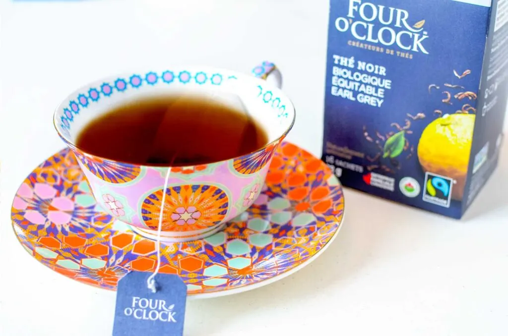 an elegant set up of a tea cup filled with brown tea, and a tea bag stil floating inside. The label is sticking out, attached to the string is a navy label for four o clock tea. It's sitting in a more modern tea cup with geometric pink, red, purple, and green shapes. Behind it is the box for four oclock earl grey tea.