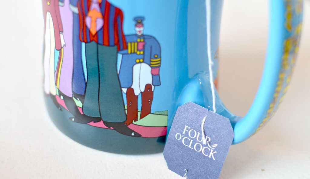 a close up of the bottom of a blue mug with cartoon people on it sit on a table. The focus was to show the tea bag label hanging down, which says, four o'clock