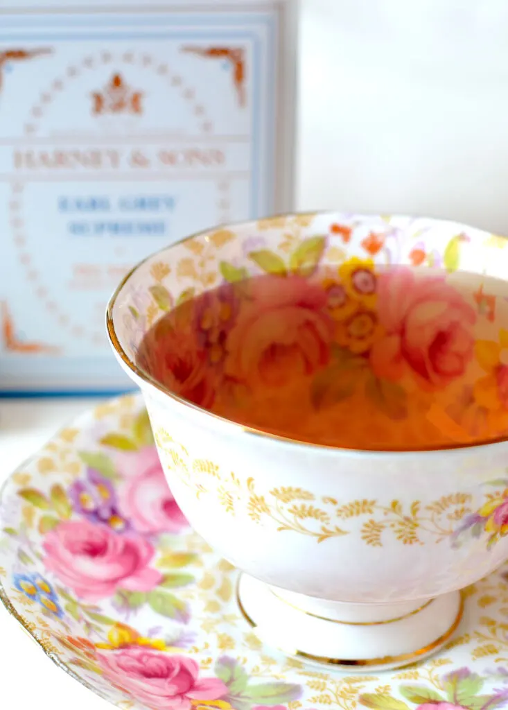 a beautiful white floral teacup with pink, gold, and yellow accents sits at the front of the picture. It's filled to the brum with a deep reddish brown brew. In the background is a blurred tin for harney and sons surpreme earl grey tea
