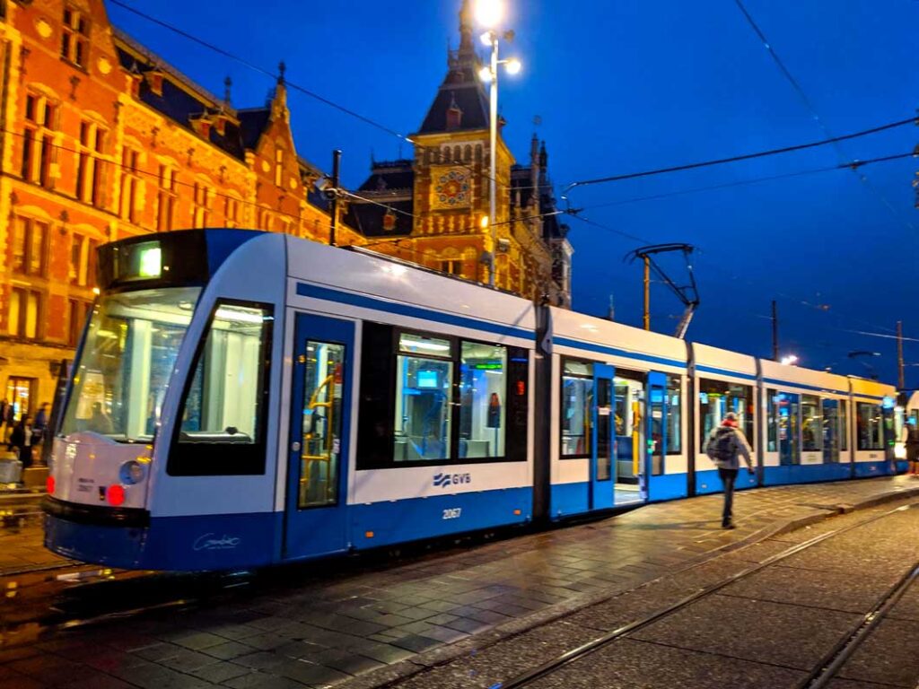 a blurry white and blue tram as it rushes forward in the photo on the streets of amsterdam at night. The night sky is a beautiful royal blue. The red brick building behind the tram is lit up with yellow lights, turning it orange. And yellow lights light up the stone streets adding a beautiful tri tone to the image of orange, yellow and blue. 