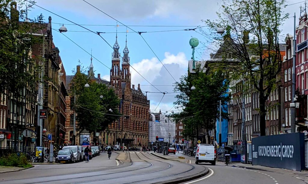 the street of amsterdam, you can see the tram tracks in the middle of the street and the wires above. There a twin spired red brick building at the end of the street. 