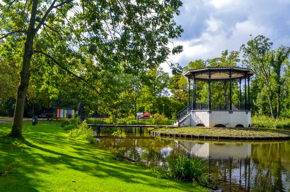 vondelpark in amsterdam. lush green grass, with trees and the start of a pond. There's a tall white gazebo just in the middle of the pond, needed a bridge to get there. 