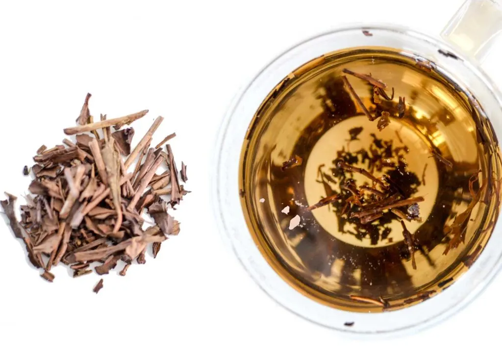 Hojicha roasted japanese green tea. A birds eye view of the dried, branch like, tea leaves. And a brewed hojicha tea in a clear double walled glass mug. It's caramel brown colour when brewed