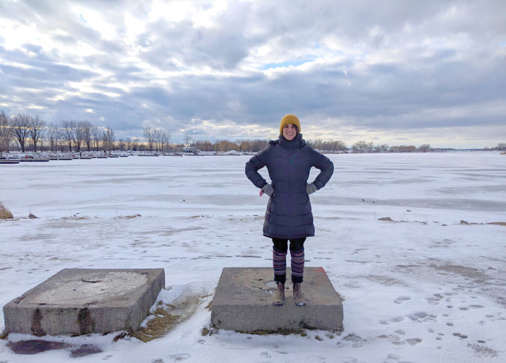 a woman stands up tall with her hands on her hips presenting her outfit. She's standing on a cinder block in the middle of a snow field park with the icy waters behind her. She's wearing blundstone boots, leg warmers, a long insulated black puffer acket, gloves, a plaid scarf, and a mustard yellow hat