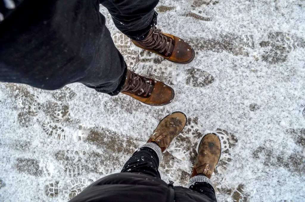 look from above of two sets of legs, a man and a woman wearing winter boots. The woman is wearing brown classic Blundstones with socks pulled up.  There's a light layer of snow on the ground, and you can see footprints that have walked by  on it already.