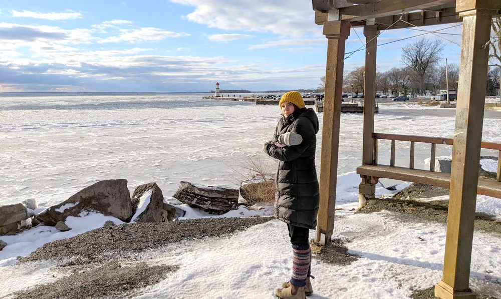 a woman pretends to be cold standing outside in Montreal winters. She has her arms wrapped around herself. She's wearing blundstone boots, corduroys, leg warmers, gloves, a scarf and a hat, in an insulated North Face jacket. Behind her is a wood pergola, meant to be a place to sit in the summer to watch the lachine canal and the lighthouse far out in the distance. Now the canal is frozen over with ice and snow