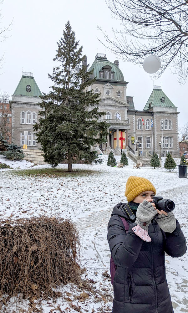 a woman stands on the corner of the phtoo, in a winter jacket, an open convertible mittens to use her camera that she's holding to take a picture of something across the street. 