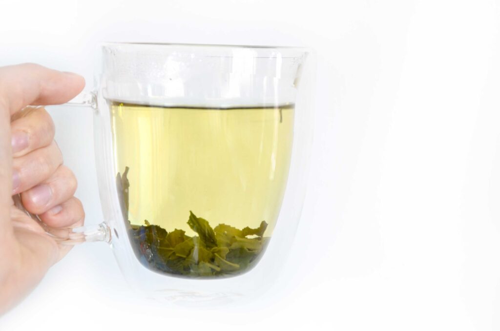 a hand holding up a clear double walled glass so you can see the brew colour: pale yellow, and the large green tea leaves that have opened up at the bottom. 