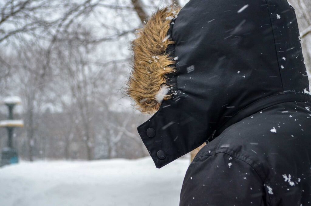 a man stands with his back at the camera, his hood is over his head with some faux fur lining it. Without the fur you could have thought that it was a black and white photo. The snow is blowing and creates a white hue to the background of the image. There's a large pile of the snow  on the ground and the trees are lined with snow. 