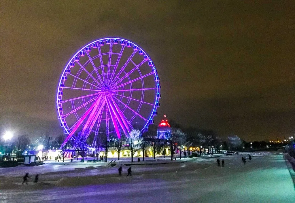 nighttime in Montreal old port but it's not quiet at all. It's hopping on Nuit Blanche with the lights on over a skating rink with people skating by, the ferris wheel behind it is lit up to a neon purple. and the lights of the city brighten the sky behind it. 