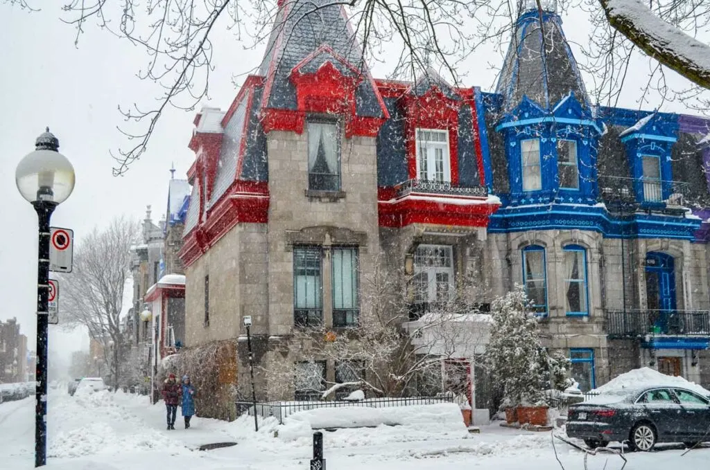 snowing in Montreal's plateau over the iconic painted ladies as they're called. They're attached three story stone homes with a triangle tower roof. They get their name from the accent colour paint on their facade, and each one is a different colour. From across the street, you can see the red and the blue house and the start of the purple one before the image cuts off. A lamp post is across from them, a car is driving by and a couple walks about to turn the corner to walk in front of them. 