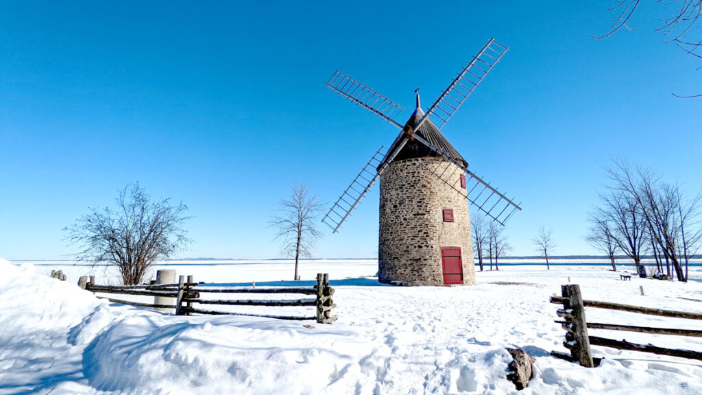 a stone windmill with a red door and covered windows in the middle of a snow field. There's a wood fence around it, that still looks like a tree