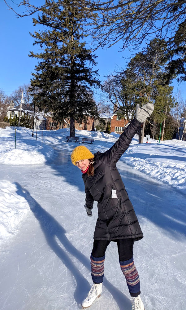 a woman spreads her arms up and down as she stopped skating to pose for the picture. She has white figure skates on with leg warmers, a north face insulated jacket, gloves, a mustard yellow hat. There's a small skate path behind her with some evergreen trees that still have leaves on them and you can see some red brick homes through the trees.