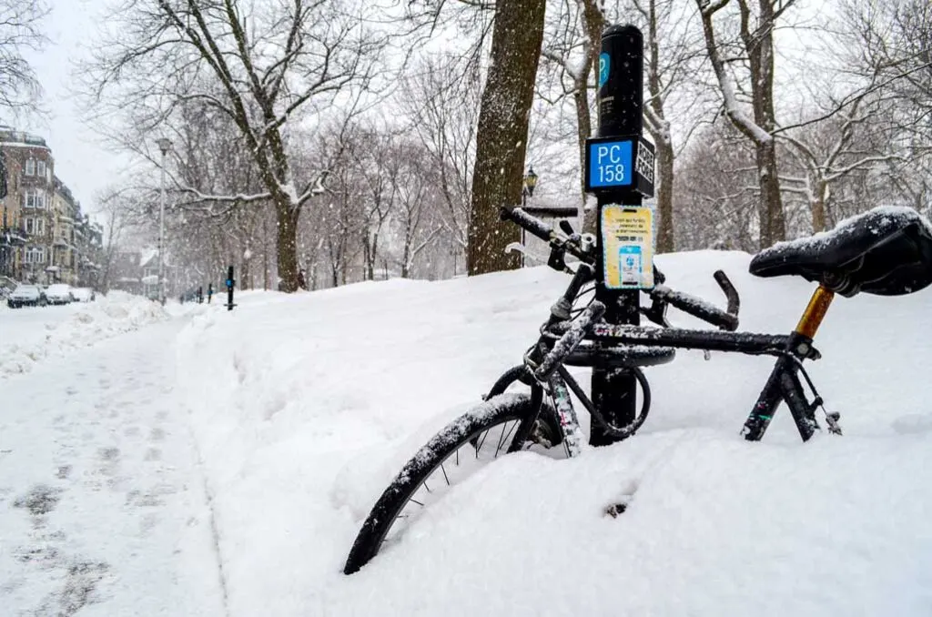 a black bike is locked up on a parking pole in Montreal. You can't see the back tire of the bike, and only half of the front tire, the rest is completely covered in a snow bank. There's is a walking path in between the snowbank. It's snowing and leaving a line of snow on the bare branches of the trees. 