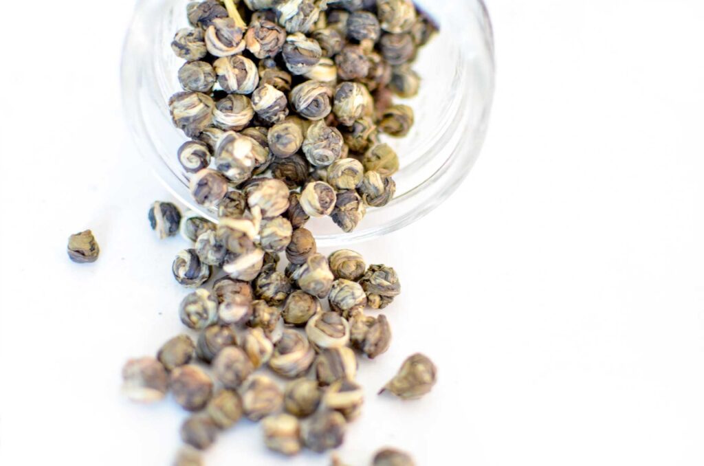 an example of chinese green tea: jasmine dragon pearls are rolled balls of green tea and the white jasmine flower. They are seen rolling out onto a while table from a glass mason jar