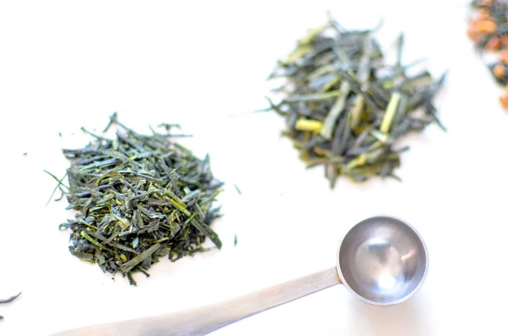 Piles of dried japanese green tea leaves in three piles on a white background with a silver teaspoon for measuring in front. Each pile of tea leaves are slightly different; the one in front are more green and smaller compared to the one next to it. The last one, which is mostly cut off in the photo, has some orange bits in it.