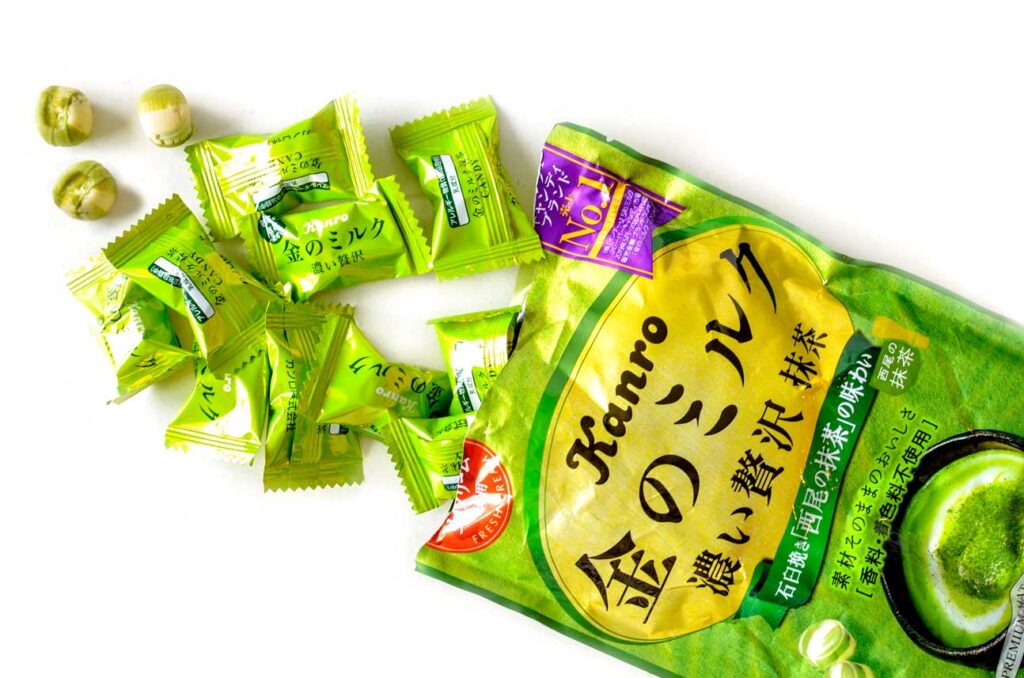 a bag of Kanro candies on a table, it's ripped open to show a bunch of smaller individual wrapped candies spilling out. Three candies are open, they are circle mixed white and green. 
