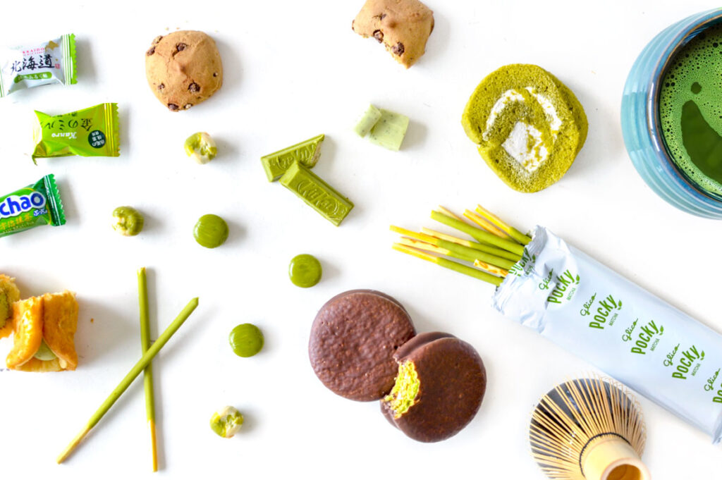 A view from above of a white table with an assortment of matcha green tea snacks. There are wrapped green candy wrappers. Green covered preztel sticks, choco pies, small hard candy green circles, green kit kat bars, chocolate chip cookies, a rolled spiral cake that's green with white cream on the inside. There's also a traditional matcha whisk and bowl that's filled with ceremonial matcha.
