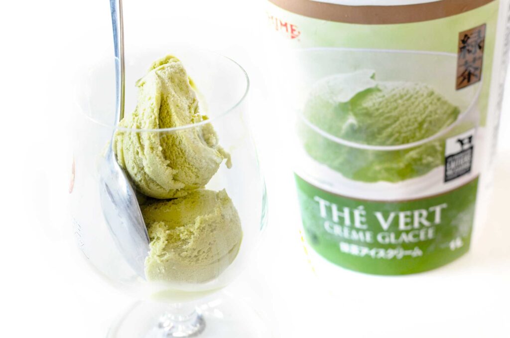 matcha green tea ice cream in a rounded glass, there are two scoops of greenish ice cream with a spoon. On the right is the ice cream container.