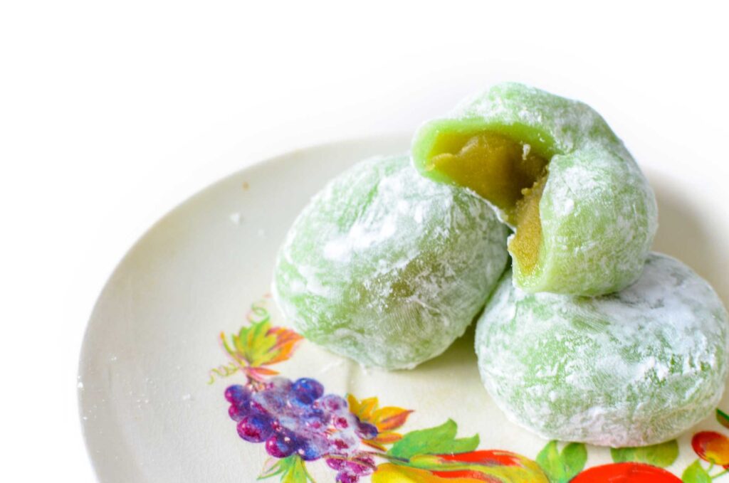 matcha-mochi-snacks-review. There are three misshapen green circular balls, with flour or powdered sugar brushed on top. There's one on top that has a triangle cut out to show the different texture inside, it's also a slightly darker more golden colour. 