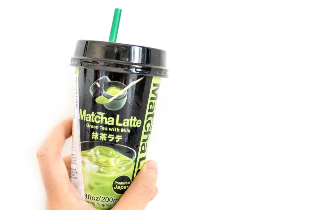 a hand is holding up a to go paper cup against a white background. The  label shows the green drink with ice in a glass, It has text in japanese, but the largest text is Matcha Latte, in smaller underneath it says Green tea iwth Milk. And above that, there's a traditional matcha powder in a tin and some on a bamboo spoon. There's a green straw sticking out the top of the lid. 