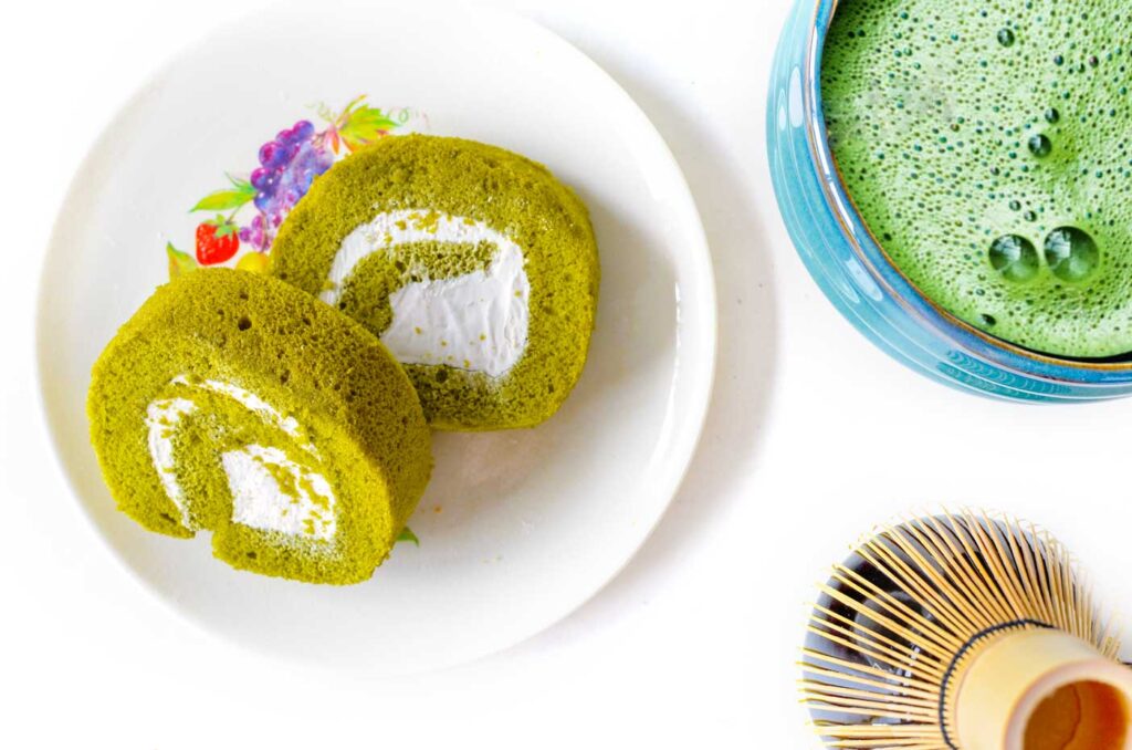 Two delicious matcha roll spiral cake on a plate. They are yellowish green with white cream filling to create the spiral. Next to the plate, half cut off are bamboo whisks and a ceremonial cup filled with green tea matcha that's still bubbly from being frothed. 