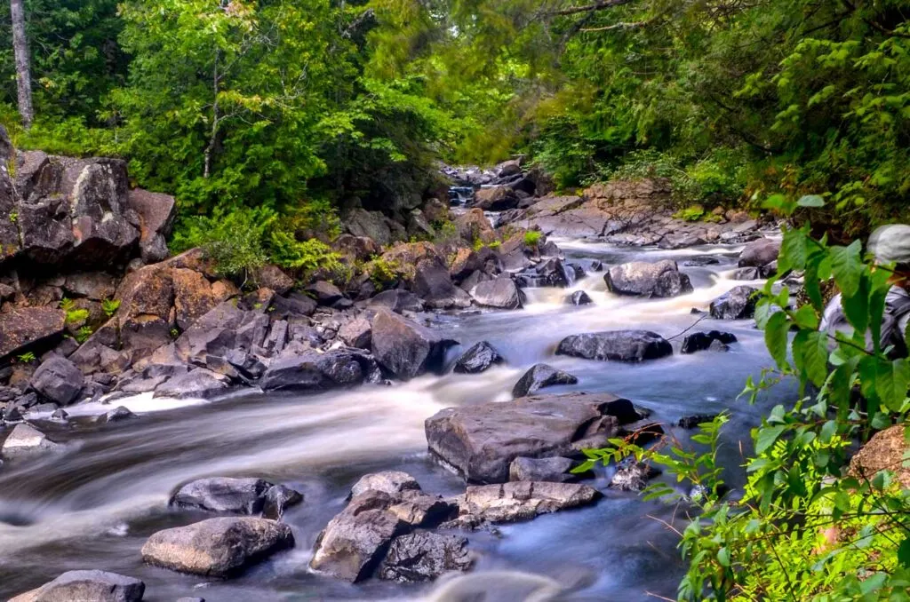 best quebec national parks near montreal. a long exposure photograph to show the smoothness of the rushing water through the rocks in the river. The river runs between green trees on either side. 