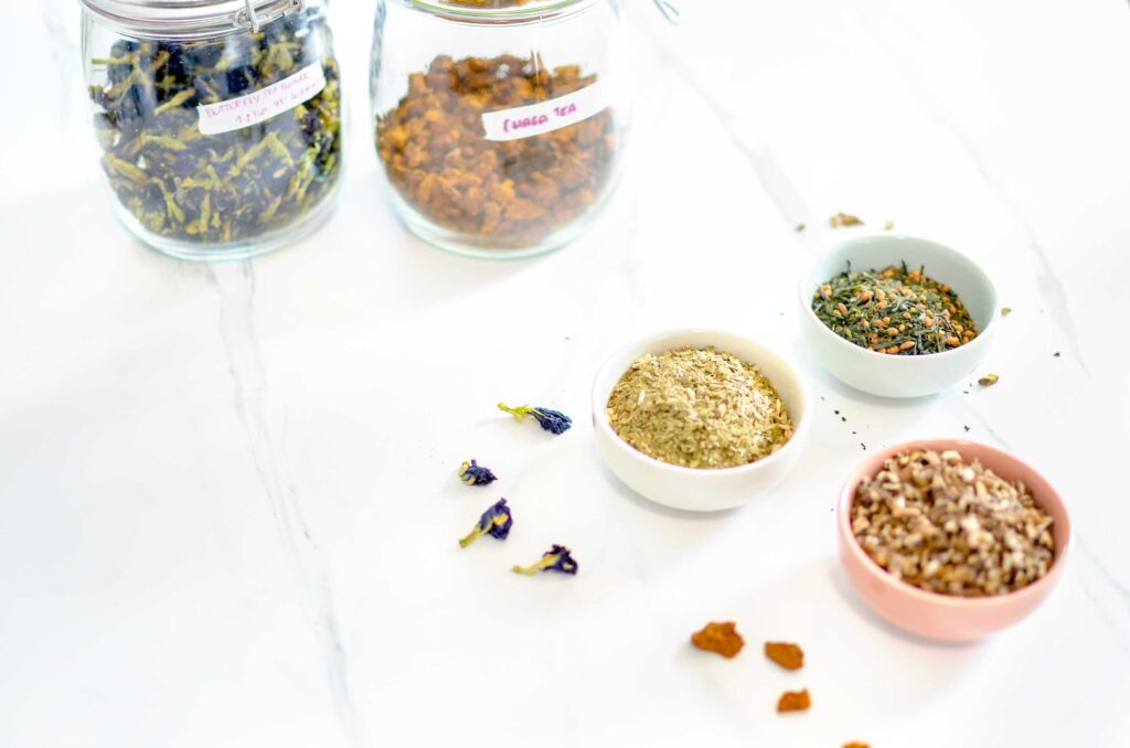 three pinch bowls and two jars are on a white marbled counter top. the jar has labels, the top left says butterfly pea flower with brewing instructions it has dried blue and green flowers. The one next to it says chaga, it has half a jar of orange looking rocks. 

the jars in front of them have three different types of tea. The top on has thin green strips wtih roasted rice. The one below has hay coloured bits. And finally the bottom one has small bits of bark. 